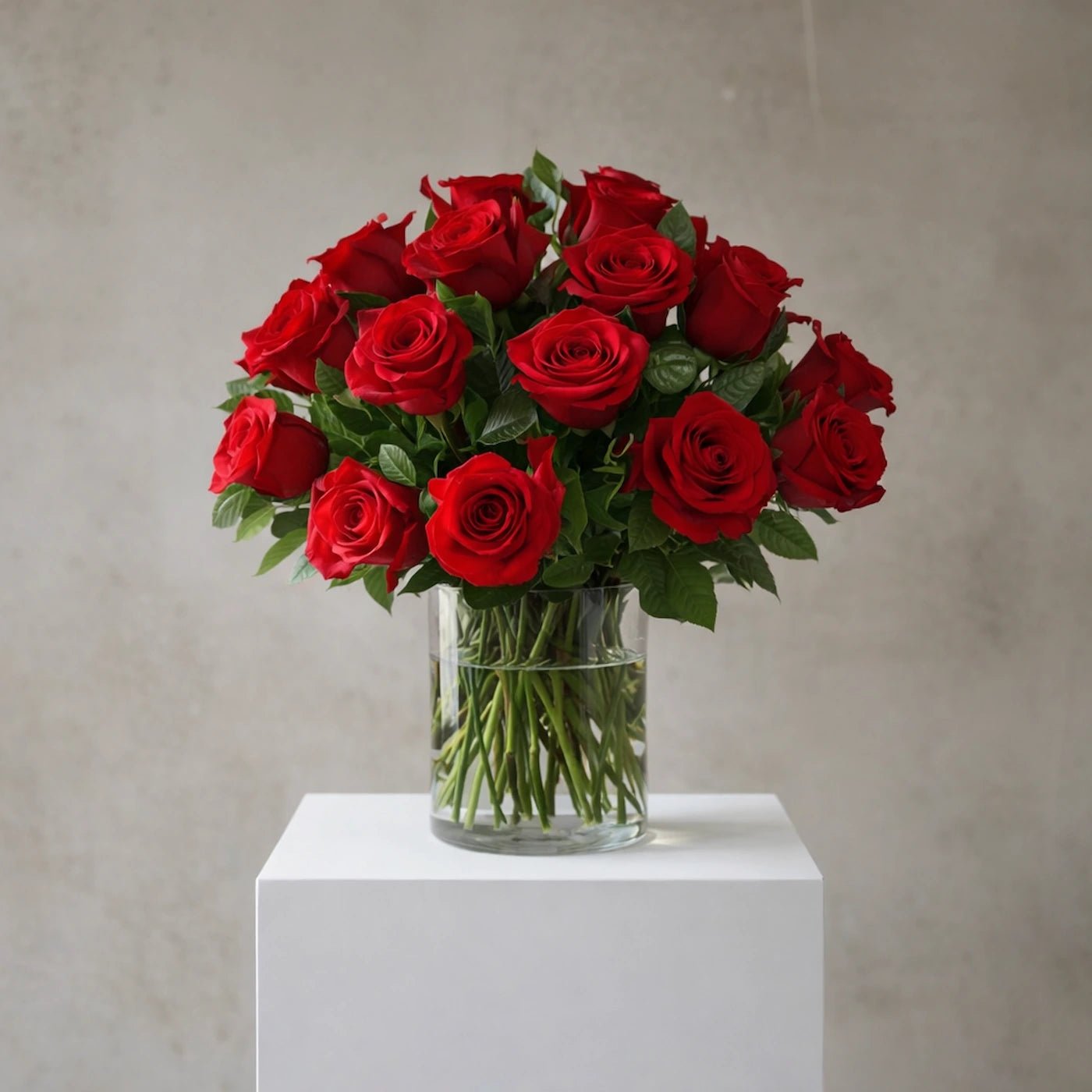 Bouquet of red roses in a clear vase on a white pedestal against a neutral background