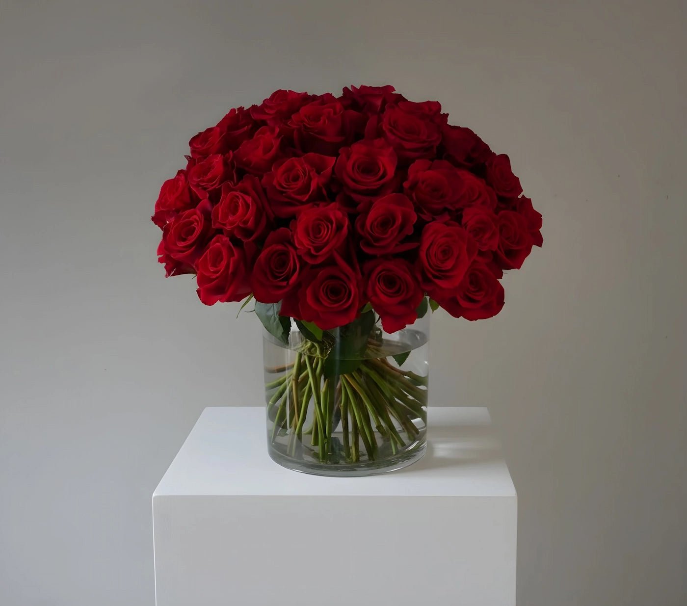 Bouquet of red roses in a clear vase on a white pedestal against a plain background