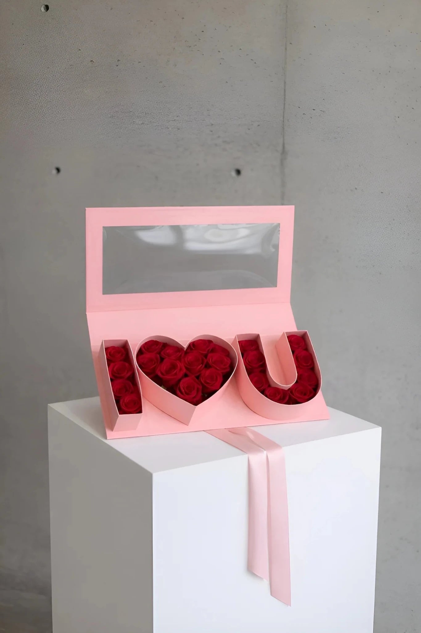 Pink jewelry box with 'I U' letters filled with red roses on a white pedestal against a gray background