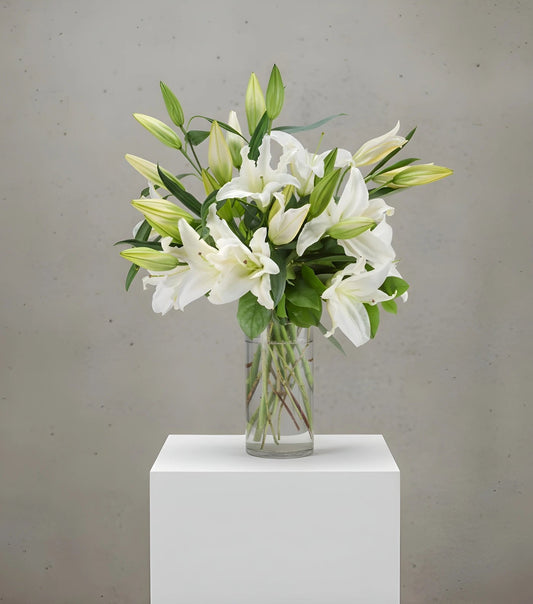 Bouquet of white lilies in a clear vase on a white pedestal against a gray background