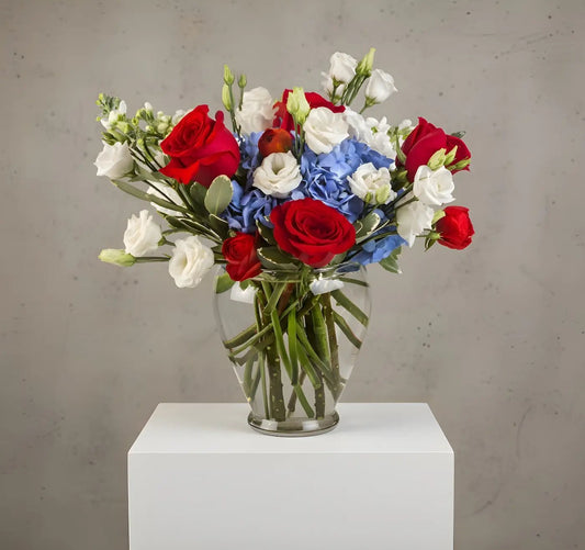 Bouquet of red, white, and blue flowers in a clear vase on a white pedestal against a gray background