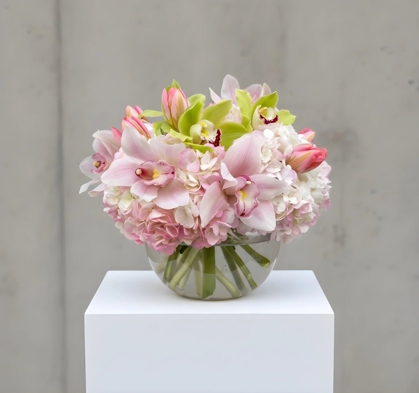 Bouquet of pink and white flowers in a clear vase on a white pedestal against a gray background