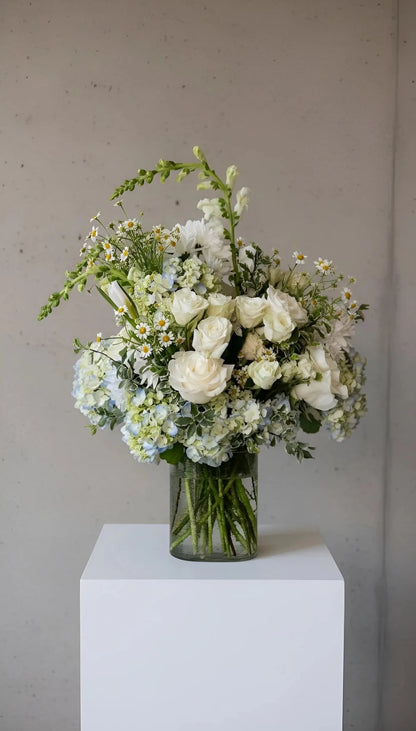 Bouquet of white and green flowers in a clear vase on a white pedestal against a plain background