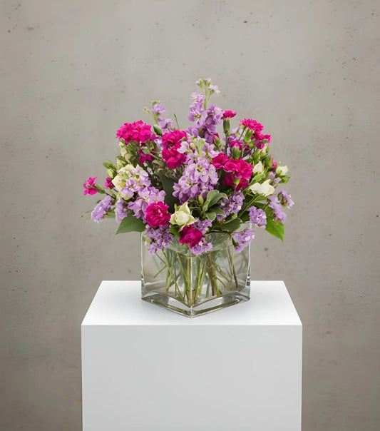 Bouquet of pink, purple, and white flowers in a clear glass vase on a white pedestal against a gray background